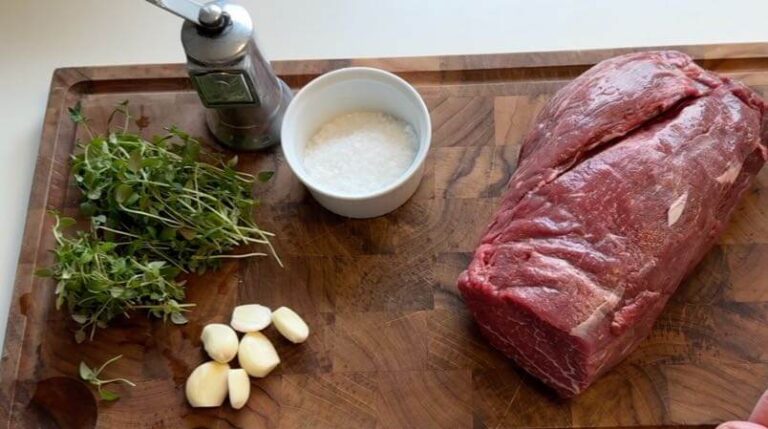 Ingredienser til sous vide oksemørbrad: Et skærebræt med frisk timian, hvidløg, flagesalt, solsikkeolie og en rå oksemørbrad – alle klar til sous vide-tilberedning.